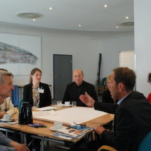 Teilnehmer sitzen diskutieren am Arbeitsgruppentisch und hören dem gestikulierenden Sprecher zu. Alle schauen freundlich drein.