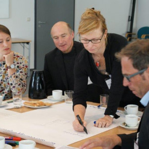 An einem Arbeitsgruppentisch steht eine Teilnehmerin und schreiben mit einem schwarzen Stift einen Beitrag auf Papier. Die anderen Teilnehmer schauen interessiertzu.