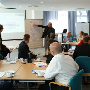 Ein Teilnehmer erklärt Gruppenarbeitsergebnisse zeigend am Flipchart bestückt mit einer handschriftlichen Grafik. Die anderen Teilnehmer sitzen an den Gruppenarbeitstischen und hören zu.