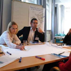Teilnehmer sitzen an einem Gruppenarbeitstisch. Eine Teilnehmerin schreibt die ausgesprochenen Gedanken eines anderen Teilnehmers auf Papier. Andere Teilnehmer folgen dem interessiert.