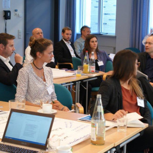 Der Blick geht hinein in den Arbeitsgruppenraum, wo die Teilnehmer an den Arbeitsgruppentischen sitzen. Ein Teilnehmer erzählt etwas. Andere Teilnehmer hören zu. Eine Teilnehmerin dreht sich dem Erzählenden zu.