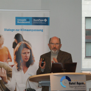 Vortragender steht am Rednerpult vor einem aufgeklappten Laptop und trägt gestikulierend vor. Vorne auf dem Stehpult steht der Name des Tagungszentrums Aquino und im Hintergrund steht das Rollup Dialoge der Klimaanpassung und man sieht rechts durch ein Fenster hinaus auf eine Häuserfassade.