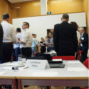 Teilnehemende stehen in lockeren Gesprächsgruppen beieinander. Sie tragen vorwiegend weiße Hemden und blaue Hosen. Im Vordergrund stehen ein Tischschild mit der Bezeichnung Landkreis Hiese.