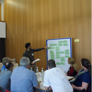 Ein Teilnehmer zeigt auf eine Präsentationswand mit grünen Moderationskarten. Im Vordergrund sitzen Teilnehmende am Gruppenarbeitstisch und hören zu.