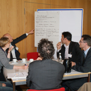 Teilnehmer sitzen am Gruppenarbeitstisch. Ein Teilnehmer zeigt mit ausgestrecktem Arm auf beschriftetes Papier an Moderationswand im Hintergrund.