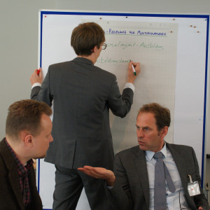 Teilnehmer schreibt auf Papier an Moderationswand. Im Vordergrund sitzen zwei Teilnehmer im Gespräch.