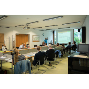 Workshopteilnehmende sitzen in einem hellen Raum an einer rechtigen Tischformation. Der Blick geht hinaus zum Fenster. Vor dem Fenster steht die Präsentationsleinwand. Der Vortragende zeigt auf seine Präsentation. Er trägt ein hellblaues Shirt.