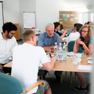 Planspiel: Teilnehmende sitzen an Gruppenarbeitstischen und denken nach. Im Vordergrund schreibt ein Teilnehmer im weissen T-Shirt etwas auf eine hellblaue Moderationskarte.
