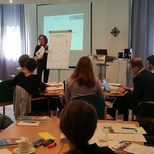Vortragende steht am Flipchart und erklärt Ergebnisse. Teilnehmende sitzen an Gruppenarbeitstischen und hören zu. Im Hintergrund sieht man das Sonnenlicht durch die zugezogenen Gardinen durchscheinen.