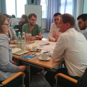 Teilnehmende sitzen im angeregten Gespräch miteinander am Gruppenarbeitstisch. Auf dem Tisch liegen Materialien wie Papier, Stifte, bunte Blätter. Es stehen Getränkeflaschen mit Wasser auf den Tischen sowie Tassen und Gläser.