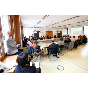 Workshopgruppenraum Ambiente im Umweltbundesamt: Teilnehmende sitzen im Rechteck an Tischen und hören dem Vortragenden zu. Der Raum ist in hellen Farben gestaltet. Manche Teilnehmende haben sich auf Stühlen und Fensterbänke verteilt.