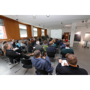 Teilnehmende sitzen in lockeren Stuhlreihen vor einer Präsentionsfläche  im Ausstellungsraum des Umweltbundesamtes. Links und recht neben der Projektsflächen stehen die Vortragenden. Ein Flipchart und eine Stellwand stehen daneben aufgereiht.