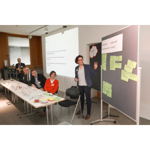 Eine Teilnehmerin präsentiert Gruppenarbeitsergebnisse an einer Pinwand. Die Ergebnisse stehen stehen auf hellgrünen Moderationskarten geschrieben. Im Hintergrund ist eine Präsentation eingeblendet. Davor steht ein Teilnehmender am Stehpult.