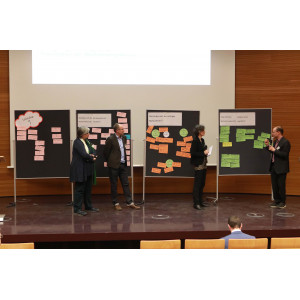 Abschlusspräsentation auf dem Podium im Hörsaal. Auf dem Podium stehen 4 Stellwände nebeneinander. Auf den Stellwänden befinden sich die Workshopergebnisse auf farbigen Moderationskarten geschrieben angepinnt. Und die jeweiligen Moderatorinnen und Moderatoren erläutern die Ergebnisse. Zwei Personen halten ein Mikrofon in der Hand.