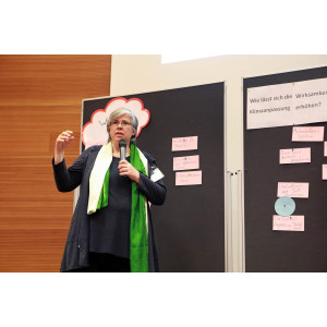 Teilnehmerin erläutert vor einer dunklen Pinwand mit hellrosa Moderationskarten die Workshopergebnisse. Sie hält ein Mikrofon in der Hand und trägt einen grün beigen Seidenschal.