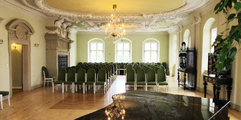 Barocksaal mit Stuckdecke und Holzparkettboden, im Vordergrund steht ein schwarzer Flügel, der Raum ist bestuhlt