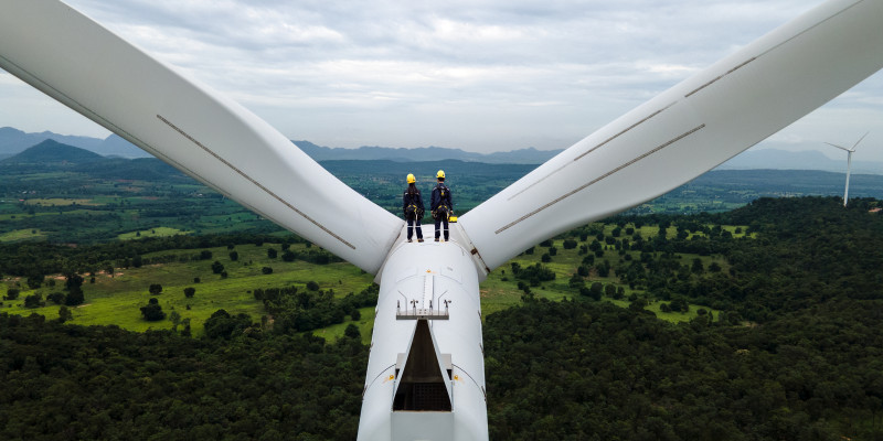 Zwei Menschen i9n Arbeitskleidung stehen auf einer Winkraftanlage und schauen in einer bewaldete Landschaft.