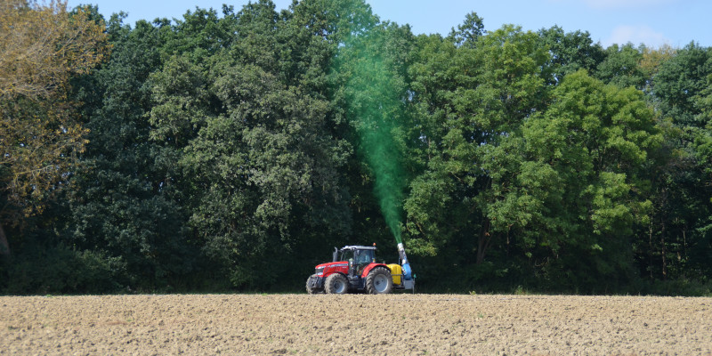 Das Bild zeigt einen Traktor an einem Waldrand, der mit Hilfe einer Sprühkanone Biozide in Richtung der Bäume ausbringt.