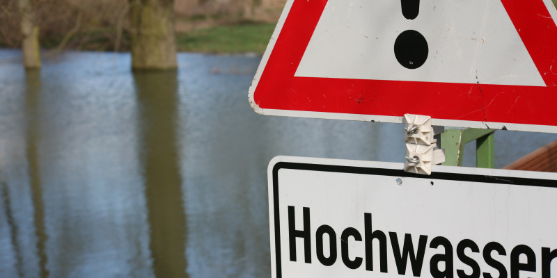 Foto: Ein überschwemmtes Gebiet während eines Hochwasserereignisses. Im Vordergrund steht ein „Achtung Hochwasser“ Schild