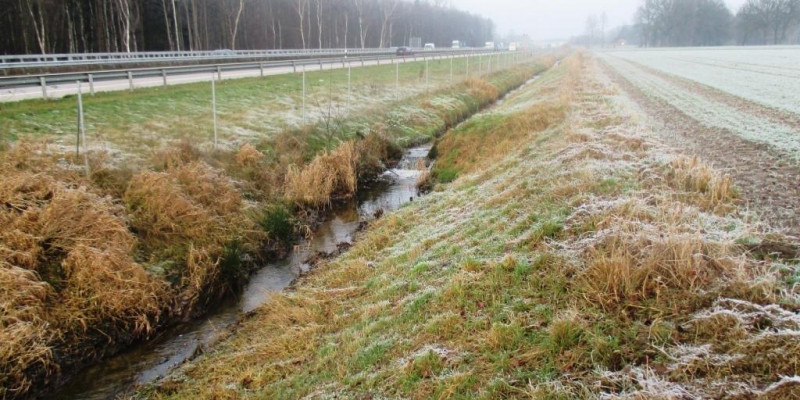 Foto: Der begradigte Schierenbach im V-Profil eingeengt zwischen Autobahn und Ackerfläche. Die Ufer sind bis auf kurze Gräser frei von Vegetation.