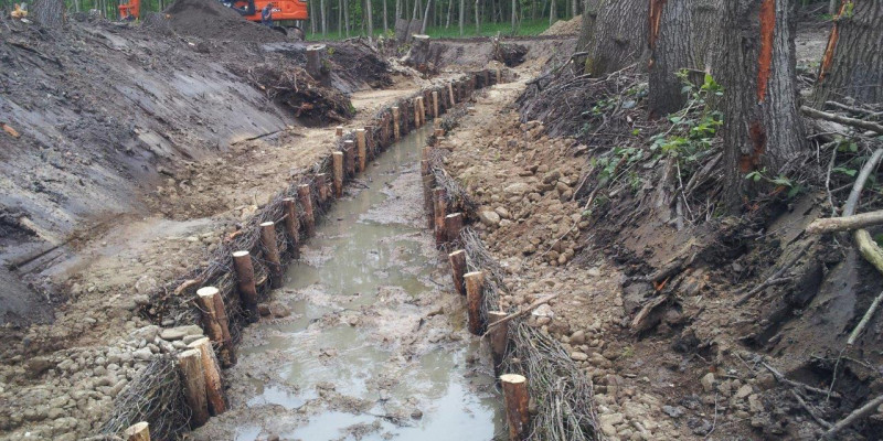 Foto: Einbau einer naturnahen Uferbefestigung aus Faschinenwalzen, Kieshinterfütterung und Wurzelstöcken am Schierenbach. Die Ufer sind noch frei von Vegetation. Im Hintergrund arbeitet ein Bagger.