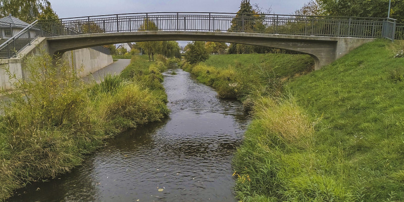 Foto: Eine Brücke über die Helme in Sundhausen. Links am Ufer ist eine Hochwasserschutzmauer zu sehen. Das rechte Ufer ist aufgeweitet und naturnah gestaltet. Am Gewässer wachsen Gräser und Sträucher.