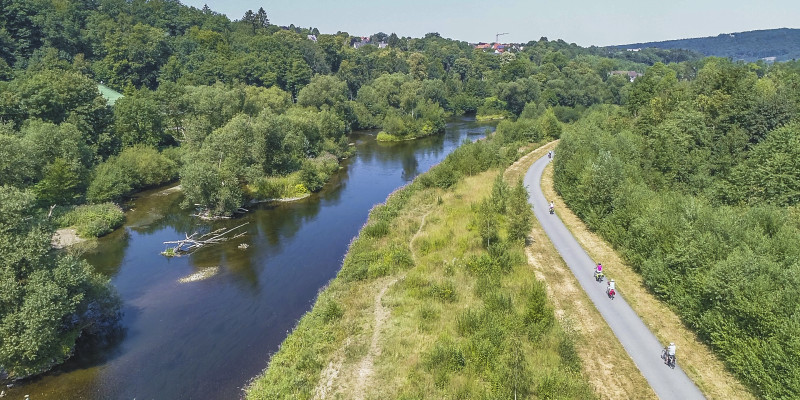 Foto: Der Fluss Ruhr bei Arnsberg mit zahlreichen Inseln, Kiesbänken, Totholz und dichtem Bewuchs. Etwas abseits des renaturierten Flusses verläuft ein befestigter Weg auf dem mehrere Personen auf Rädern unterwegs sind.