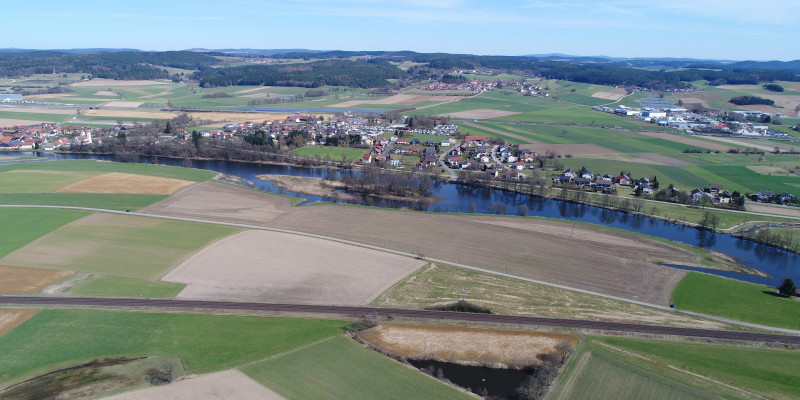 Luftbildaufnahme des Flusses Naab mit Grünland- und Ackerflächen, die bis an die Gewässerkante reichen. Parallel zum Fluss verlaufen auf beiden Seiten Verkehrswege.