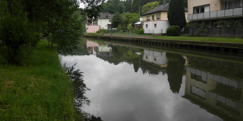 Foto: Ein begradigter Fluss mit Ufersicherung und einseitig mit Häusern, die bis auf wenige Meter an den Gewässerrand reichen.