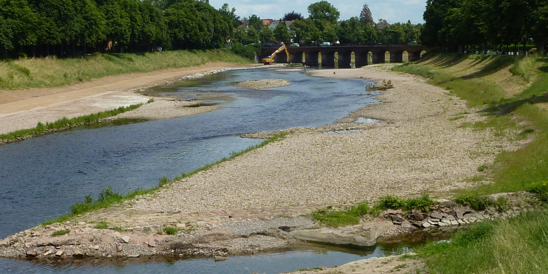 Foto: Ein renaturierter Gewässerabschnitt mit ausladenden Kiesbänken. Noch sind die Ufer vegetationsfrei. Im Hintergrund steht ein Bagger an einer Brücke. 