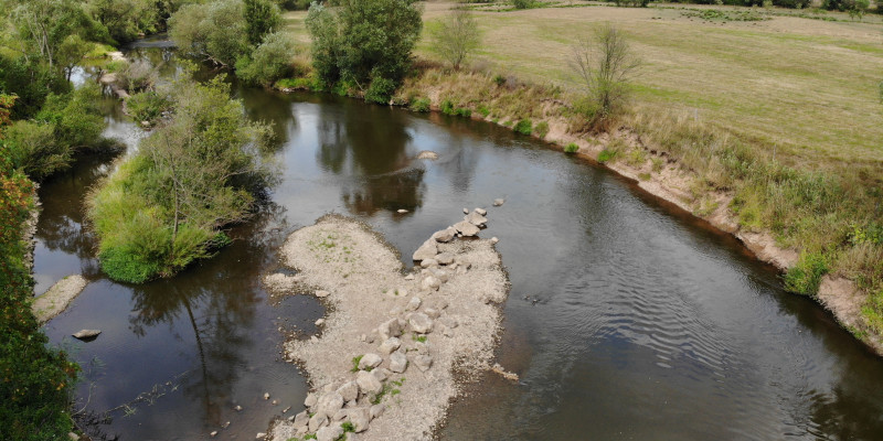 Luftbild an der Fulda. Eine Reihe von großen Steinen sind als Strömungslenker schräg in das Gewässerbett eingebaut. Durch die Ableitung der Strömung beginnt das Ufer auf der gegenüberliegenden Seite zu erodieren.
