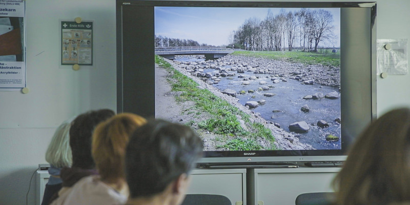 Foto: In einem Konferenzzimmer schauen mehrere Menschen auf einen Bildschirm, auf dem ein Bild einer Renaturierung gezeigt wird. 