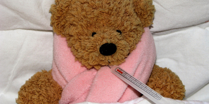Teddybär liegt mit Schal und Fieberthermometer im Bett