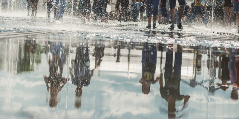 Das Bild zeigt eine Wasserfläche in einer Stadt, offensichtlich ein Brunnen. Es laufen mehrere Menschen über die Wasserfläche und spiegeln sich darin.