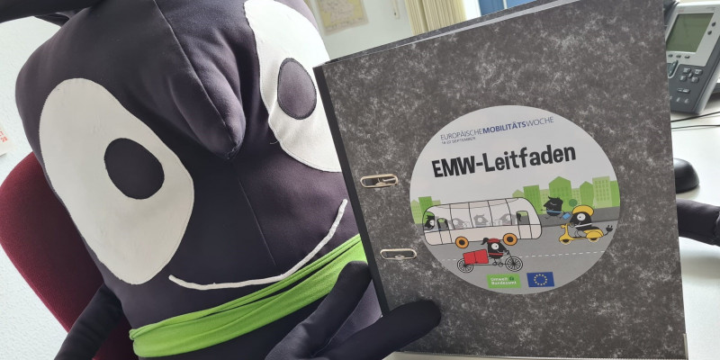 Stoffpuppe des EMW-Maskottchens hält den neuen EMW-Leitfadenordner hoch.