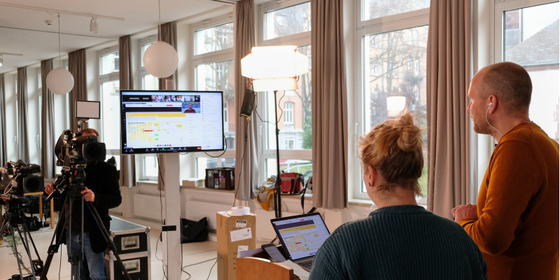 Zwei Moderatoren gucken in die Kamera und auf einen Bildschirm, während Sie das Netzwerktreffen moderieren.