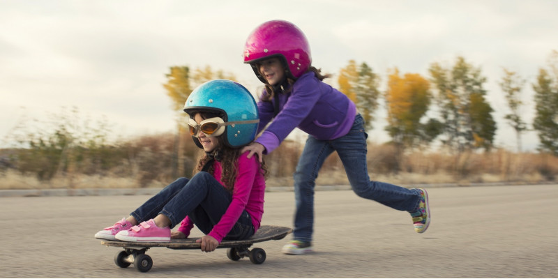 Ein Kind mir Helm schiebt ein anderes Kind mit Helm auf einem Skateboard. 
