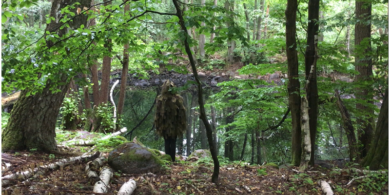 Fotografie eines Menschen, der mit Naturmaterialien verkleidet ist und im Wald vor einem Teich steht