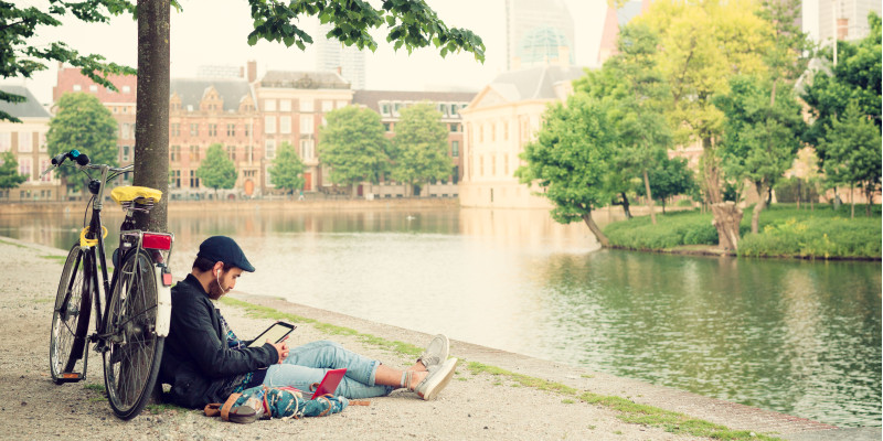 Ein junger Mann sitzt in der Stadt an einem Fluss unter einem Baum und schaut auf sein Tablet mit Kopfhörern im Ohr. Am Baum lehnt sein Fahrrad.