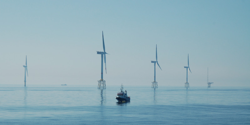 Das Bild zeigt im Meer stehende Windräder eines Offshore-Windparks und ein dazwischen schwimmendes Schiff.