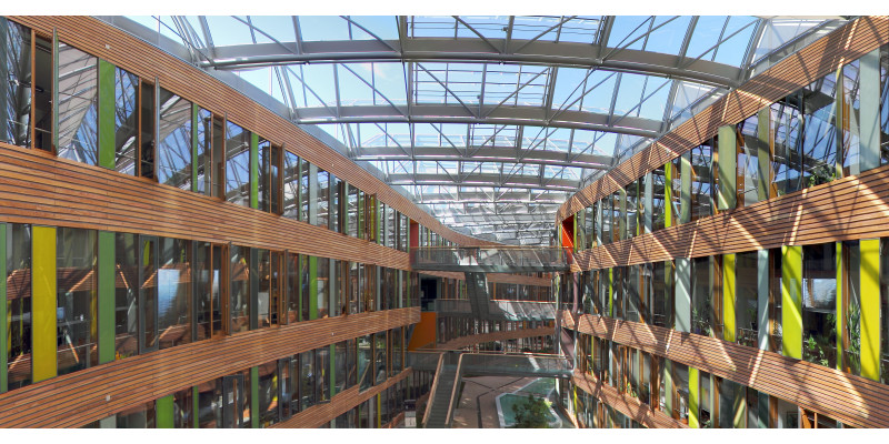 Blick in den mit Glas überdachten Innenhof, rechts und links die Innenfassaden mit Holzvertäfelungen und Glasscheiben in Grüntönen, im Innenhof Brücken und Treppen, die die Gebäudeteile verbinden