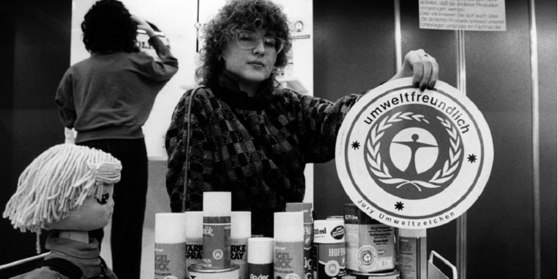 Eine Frau hält das Umweltzeichen in die Kamera, vor ihr auf einem Tisch viele Produkte, die das Umweltzeichen tragen, zum Beispiel Rasierschaum-Flaschen.