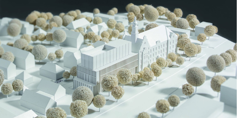 ein Architekturmodell zeigt ein historisches viergeschossiges Gebäude mit Giebel und Türmchen und einen ebenso hohen modernen Anbau inmitten einer Stadt mit Straßen, Bäumen und niedrigeren Häusern
