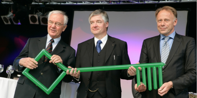 Gruppenfoto: drei Männer in Anzug und Krawatte halten einen riesigen grünen Schlüssel, im Hintergrund Sektgläser