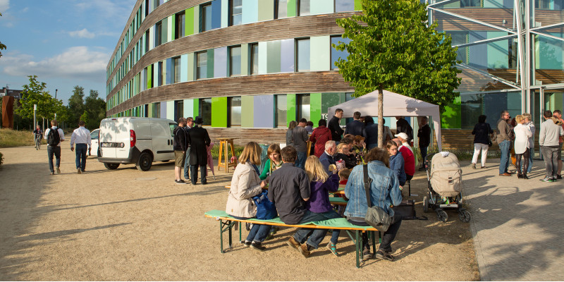 essende Menschen an Biertischen mit Bierbänken vor dem UBA Dessau-Roßlau, an einem weißen Zeltling stehen Menschen an