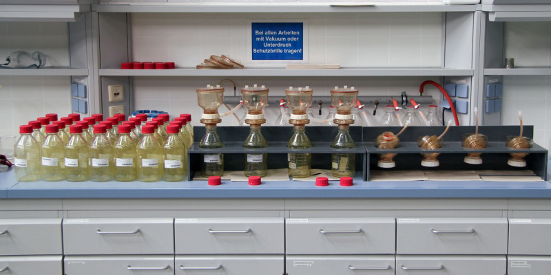 in einem Labor stehen auf einer Arbeitsplatte viele durchsichtige Gefäße mit roten Deckeln, an der Wand hängt ein Schild: Bei allen Arbeiten mit Vakuum oder Unterdruck Schutzbrille tragen!
