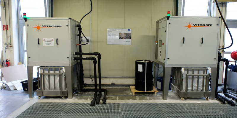 teschnische Anlage in einer Werkshalle aus zwei Boxen, aus denen Rohre in eine Gitterabdeckung im Fußboden führen