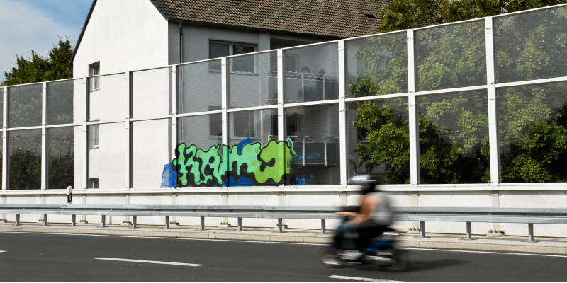 ein mehrstöckiges Wohnhaus mit Balkonen steht direkt hinter einer Lärmschutzwand einer Stadtautobahn