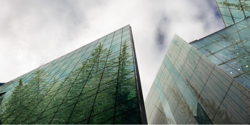in modernen Gebäuden spiegeln sich grüne Bäume in den Glasfassaden, oben ein blauer Himmel mit weißen Wolken