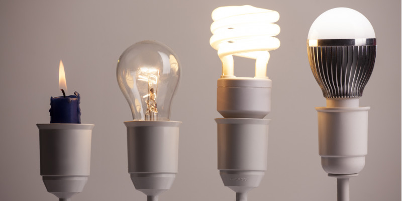 vier Lampenfassungen in einer Reihe, darin stecken (von links nach rechts): eine Kerze, eine Glühlampe, eine Energiesparlampe (Kompaktleuchtstofflampe), eine LED-Lampe